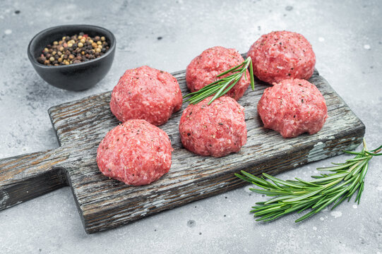 Fresh raw meatballs or frikkadel with rosemary and pepper on rustic wooden board over gray background ideal for cooking, protein and food prep themes.