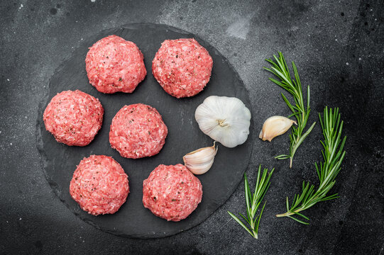 Fresh raw meatballs or Polpette with rosemary and pepper on round black slate board over dark background ideal for cooking and protein-rich themes.