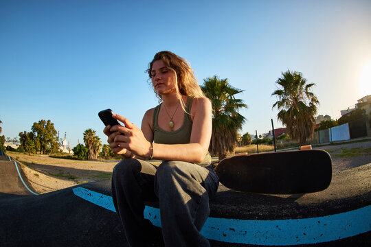 Skater checks phone while sitting near skate park at sunset