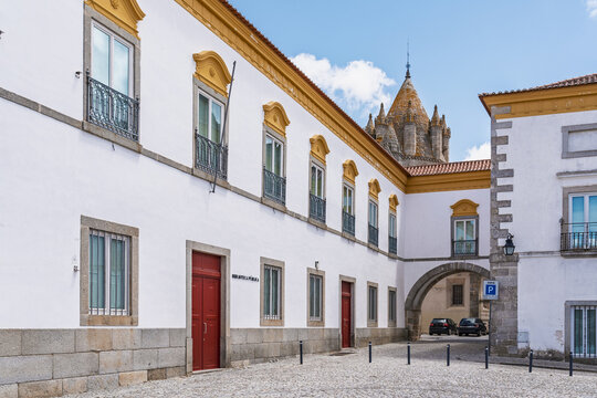 View of the historic Biblioteca Publica de Evora building with its white facade, yellow trim, and red doors, situated on a cobblestone square in Evora, Evora District, Portugal.
