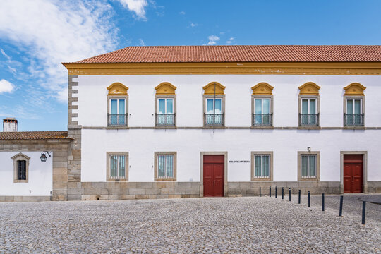 View of the historic Public Library building with its white facade, red doors, and stone base situated on a cobblestone square in Evora, Evora District, Portugal.