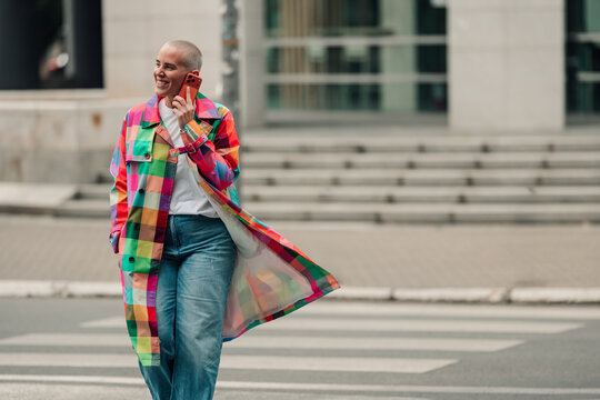 Woman with buzz cut talking on smartphone on city crosswalk, wearing a colorful plaid trench coat