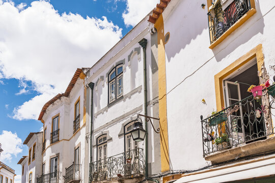 View of historic white buildings with yellow trim and ornate wrought iron balconies under a bright blue sky in Evora, Evora District, Portugal.