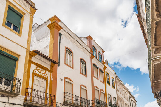 View of historic buildings with white facades and vibrant yellow and orange trim, featuring wrought iron balconies under a bright blue sky in Evora, Evora District, Portugal.