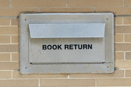 Classic Weathered Metal Book Return Box on a Tan Brick Wall, offering convenient after-hours library book drop-off service for students and readers, emphasizing education and knowledge access.