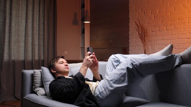 Man lying comfortably on a sofa at home, scrolling through a mobile phone, engaging with social media or online content during an evening of personal downtime and relaxation