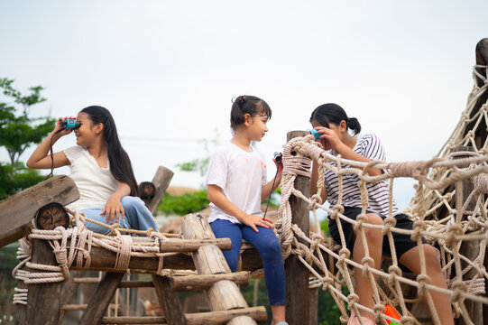 Three girls explore a playground, enjoying adventure and friendship. One uses binoculars, while another shares a drink, showcasing joyful moments.