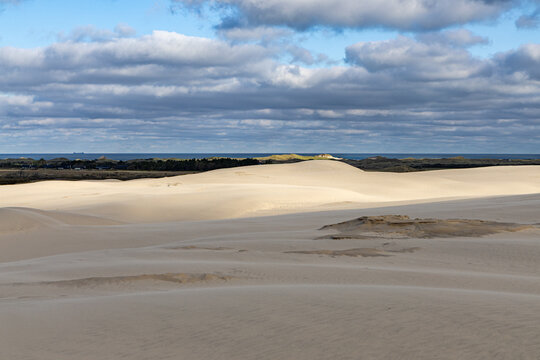 View of Rabjerg Mile sand dunes with rolling sandy hills, sparse vegetation, distant coastline, and a cloudy blue sky in Skagen, Jutland, Denmark.