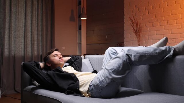 Young man lying back on a sofa with hands behind his head, relaxing and daydreaming in a cozy modern living room at night, enjoying quiet, contented alone time