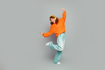 Fototapeta premium Young woman dancing in an orange sweater with yellow headphones in a photography studio, enjoying music, playful energy and casual denim streetwear