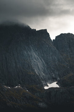 View of the craggy mountain peaks shrouded in dark, low-hanging clouds with a small patch of snow at the base in Unstad, Nordland, Norway.