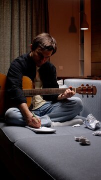Male musician on a couch composing new songs, strumming an acoustic guitar and jotting lyrics and chords in a notebook at home under warm evening light