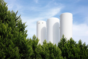 White industrial storage tanks and silos framed by green trees under blue sky © ercanerol