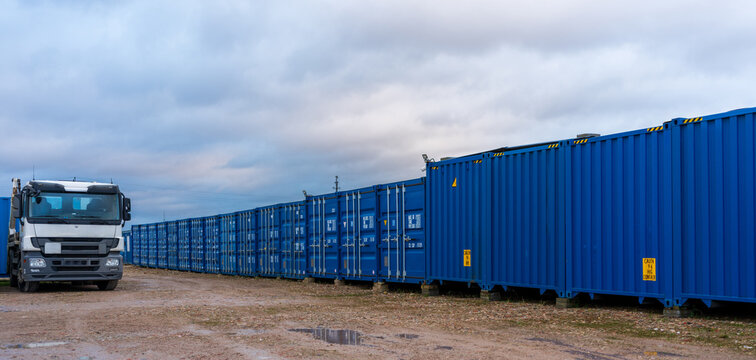 View of a white truck parked beside a long row of blue shipping containers on a dirt lot under a cloudy sky in Klaipeda, Klaipeda County, Lithuania.