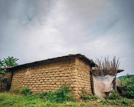 View of a traditional mud-brick house with a thatched roof and a makeshift fence made of branches and fabric in a rural landscape in Jos East, Plateau, Nigeria.