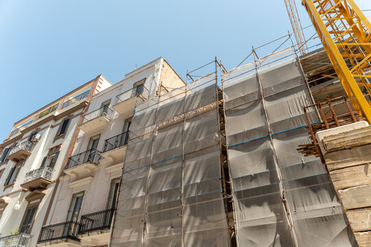 Row of old residential buildings with facades wrapped in grey mesh on scaffolding and a crane jib rising in the corner, seen from street level on a sunny day, concept of facade restoration, property 