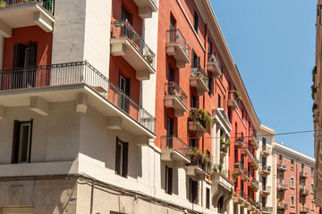 Corner apartment building with red and cream facade, wrought iron balconies and potted plants stretching along a sunny street in perspective, concept of mediterranean city living, urban architecture  © daily_creativity