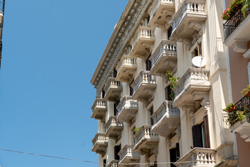 Ornate white historic building with rows of stone balconies, carved corbels and flower pots rising against a deep blue sky, low angle view with open sky on the left, concept of classic architecture,  © daily_creativity