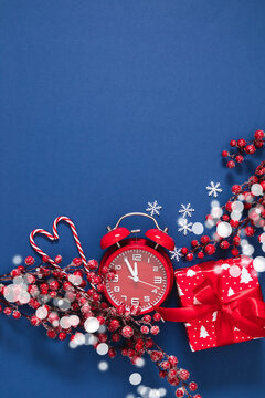 Vertical Christmas background with red alarm clock, gift box, candy canes heart, and frosty berries on blue backdrop. Festive winter holiday flat lay with copy space, snowflakes, and bokeh lights.