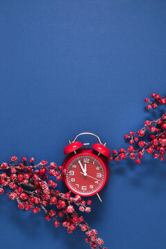 Vertical Christmas and New Year background with red vintage alarm clock showing five minutes to midnight and decorative snow covered berry branches on blue backdrop. Festive winter holiday copy space.