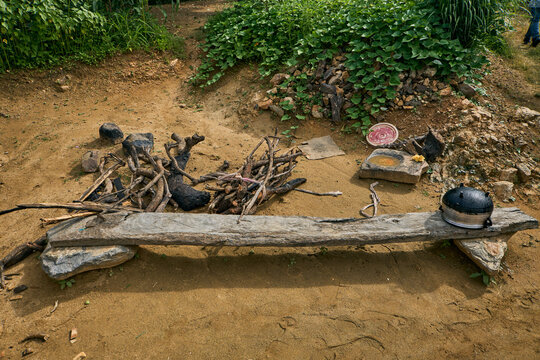 View of a rustic outdoor cooking setup with a wooden bench, firewood, a stone mortar, and a metal pot on sandy ground in Radung, Plateau, Nigeria.
