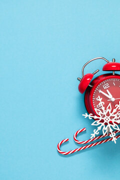 Vertical New Year background with red alarm clock showing midnight, candy canes, and white snowflake on light blue backdrop. Festive Christmas holiday side border flat lay with large copy space
