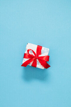 Vertical minimalist Christmas background with wrapped white gift box tied with red satin ribbon bow in center on light blue backdrop. Festive winter holiday minimalist flat lay with copy space.