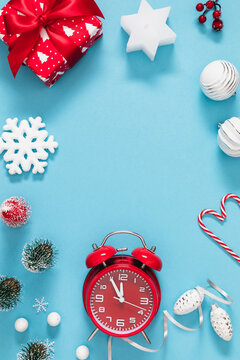 New Year vertical frame background with red alarm clock, gift box, star candle, mini trees, and snowflakes on blue backdrop. Festive Christmas holiday border flat lay with central copy space