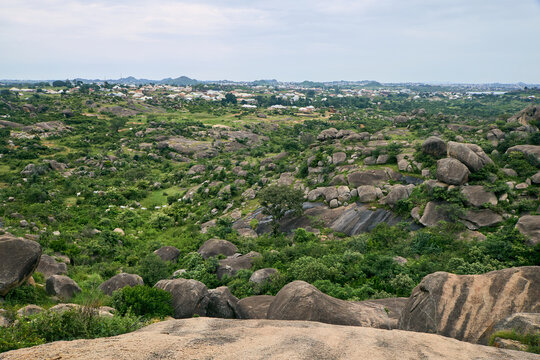 View of the rocky landscape of Shere Hills with granite boulders, lush green vegetation, and a distant town in the background in Shere Hills, Plateau, Nigeria.