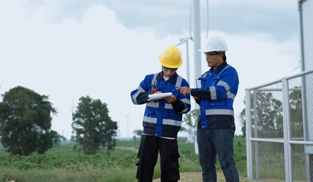 Wind turbine engineers record field inspection notes while reviewing site conditions near renewable energy equipment, working together in safety gear during an outdoor wind farm assessment.