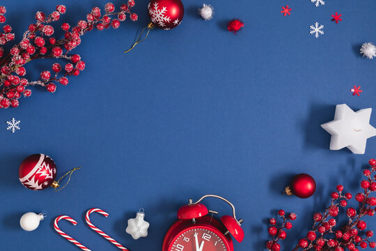 Christmas and New Year flat lay frame on dark blue background with red alarm clock, baubles, candy canes, star candle, snow covered berries, and confetti. Festive winter holiday background with copy s