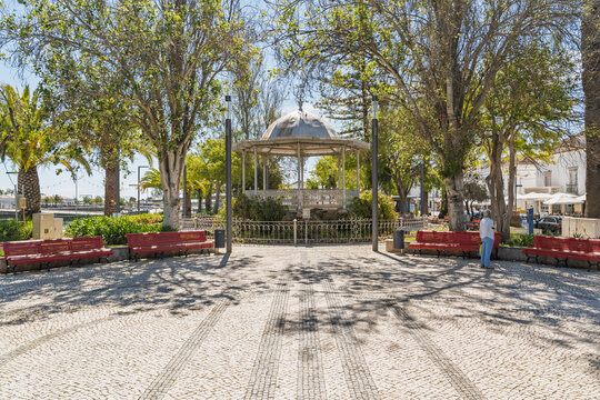 View of the historic bandstand in the Jardim do Coreto surrounded by lush trees, red benches, and traditional Portuguese cobblestone pavement in Tavira, Faro District, Portugal.