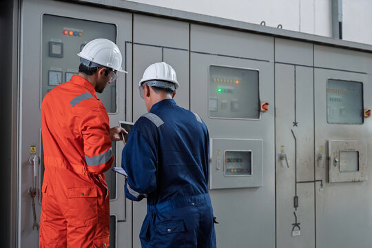 Electrical technician and engineer work together in a power distribution control room at a large industrial factory, inspecting control panels, pump systems, electrical equipment, and operating data.