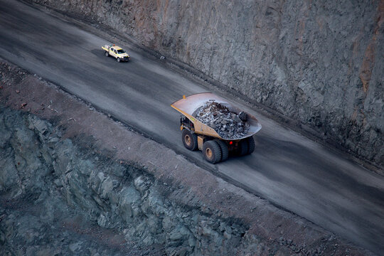 View of a large haul truck carrying rocks along a dirt road in the Super Pit mine, with a service vehicle nearby and steep rocky walls in Kalgoorlie-Boulder, Western Australia, Australia.