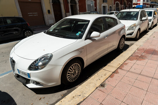 Bari, Italy - Aug 4, 2026: White Alfa Romeo Giulietta hatchback parked at a yellow painted kerb beside a red brick pavement with other white cars lined up along the sunny street