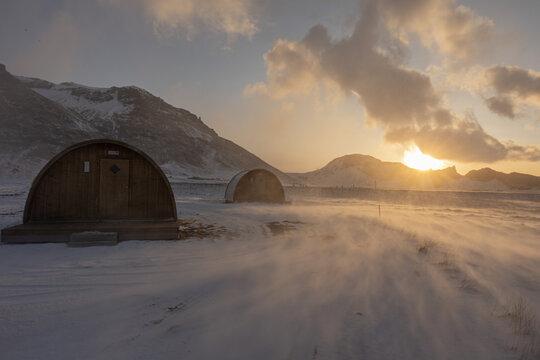 View of wooden barrel cabins in a snowy landscape during golden hour with wind blowing snow across the ground and mountains in the background in Hvolsvollur, Rangarthing eystra, Iceland.