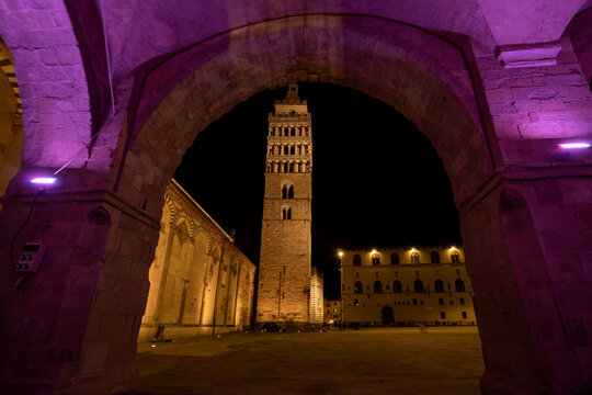 View of the historic bell tower of Pistoia Cathedral illuminated at night, framed by a stone archway with purple accent lighting, Province of Pistoia, Tuscany, Italy.