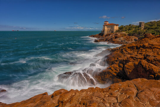 View of Castello del Boccale perched on the rugged red rocky coastline with waves crashing against the shore under a clear blue sky in Livorno, Tuscany, Italy.