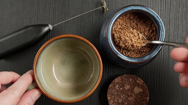 Top-down close-up of a person adding spoonfuls of instant coffee granules into a ceramic cup while preparing a hot drink at home.