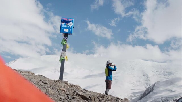 Climber drinking morning coffee beside blue mailbox on Yukhin Peak 5130m during acclimatization, snow covered Lenin Peak Ibn Sina Peak 7134m background, Pamir mountains expedition camping clip, 4K