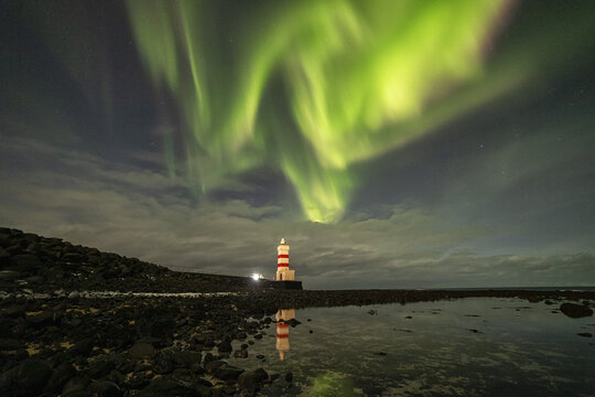View of the Gardur Lighthouse illuminated at night under a vibrant display of aurora borealis with a reflection in the rocky tide pools in Gardur, Sudurnesjabaer, Iceland.
