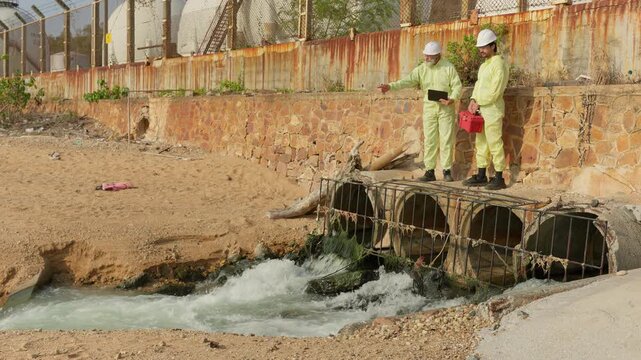 Two environmental scientists in hazmat suits inspect a sewer outfall, analyzing water quality and pollution for an environmental site assessment.