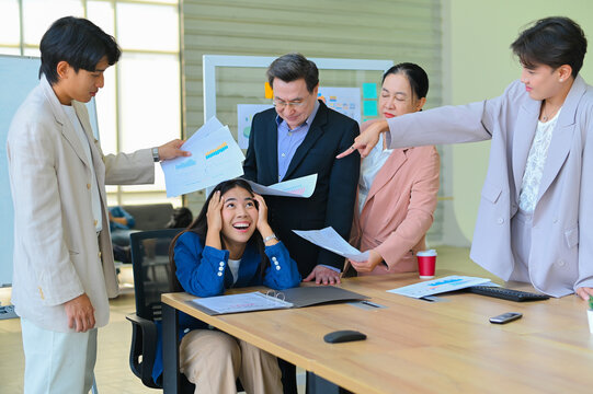 A tense meeting scene where one woman appears overwhelmed while colleagues argue and gesture with papers, showcasing workplace stress and dynamics.