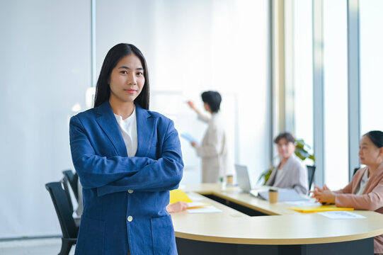 Confident businesswoman stands with arms crossed in a modern office setting, showcasing leadership and teamwork during a corporate meeting.