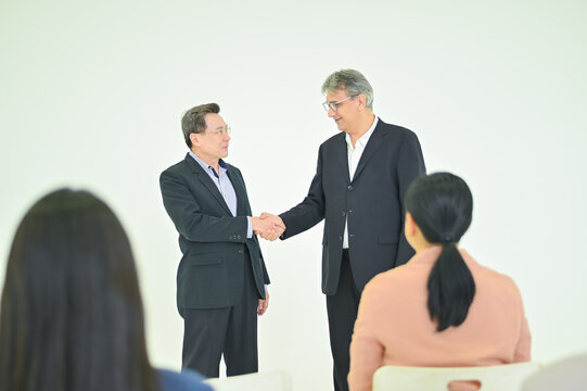 Two business professionals shake hands in a formal meeting, exemplifying partnership and collaboration in a modern workspace, with attentive colleagues observing.