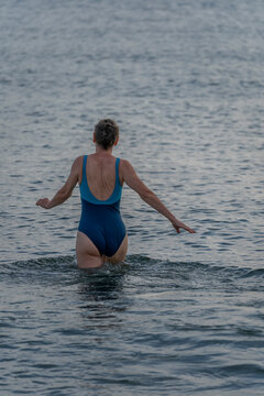 Langrune-Sur-Mer, France - 08 09 2026: A mature woman wearing a blue swimsuit from behind walks in the water to get in the sea