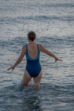 Langrune-Sur-Mer, France - 08 09 2026: A mature woman wearing a blue swimsuit from behind walks in the water to get in the sea