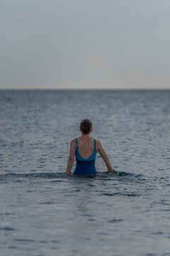Langrune-Sur-Mer, France - 08 09 2026: A mature woman wearing a blue swimsuit from behind walks in the water to get in the sea