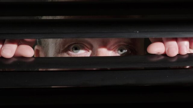 Suspicious woman looking out with astonished, frightened eyes through hole between blinds on window onto night street