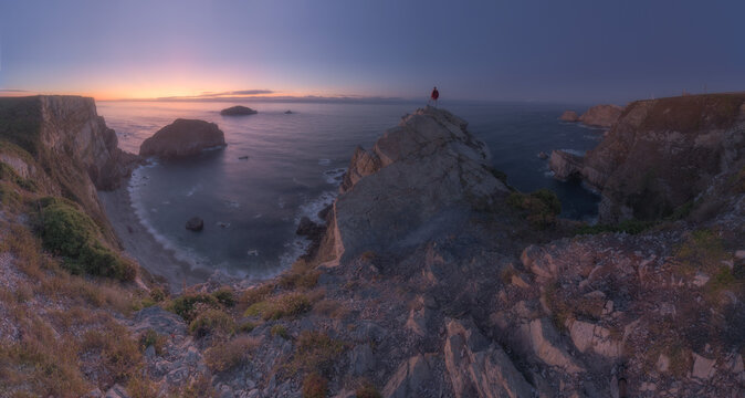 View of a person standing on a rocky cliff overlooking the ocean during a calm sunset with soft purple and orange light illuminating the coastal landscape in Spain.